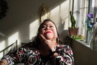 Bertha Embriz is sitting on a chair in her house beside the window. The sun is shining in, highlighting her face as she looks up to the ceiling. In the background, a statue of the Virgin Mary can be seen hanging on the wall, as well as some plants on the windowsill.