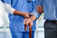 A photo of a nurse helping an older man walk with a cane in a nursing home.