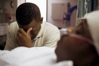 A photo of a Black man sitting beside his son in a hospital bed, covering his face.
