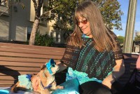 Diana McAllister is seen sitting on a park bench next to her dog, Honey. McAllister is wearing a turquoise shirt, Honey is wearing sunglasses, a visor and a skirt in the same color.