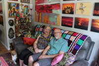 A woman and man sit on a dark gray couch covered in several colorful pillows and a crocheted blanket. Colorful artwork hangs on the walls behind and beside the couch and a small tan and white dog sits on the floor besides the couple.