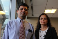 Dr. Bhavin Shah, who wears a dress shirt and tie, stands beside his wife, Sunita Kalsariya, who wears a black cardigan over a white patterned shirt. They are in an office room next to a window, and look towards the camera.